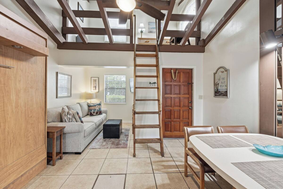 A spacious living area with a loft bedroom above, a wooden ladder, a dining table, and a sofa.