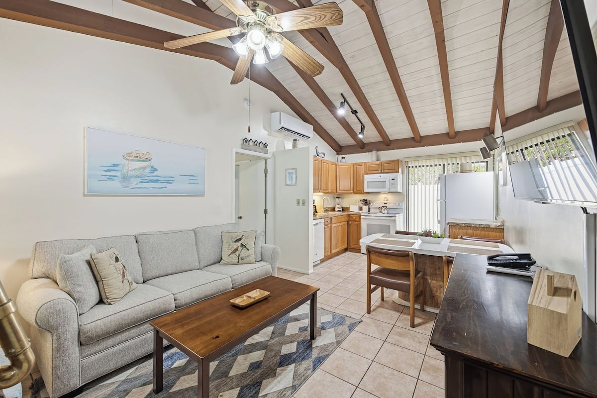 A living room with a sofa, coffee table, and a ceiling fan. There is a kitchen area with cabinets and a microwave.