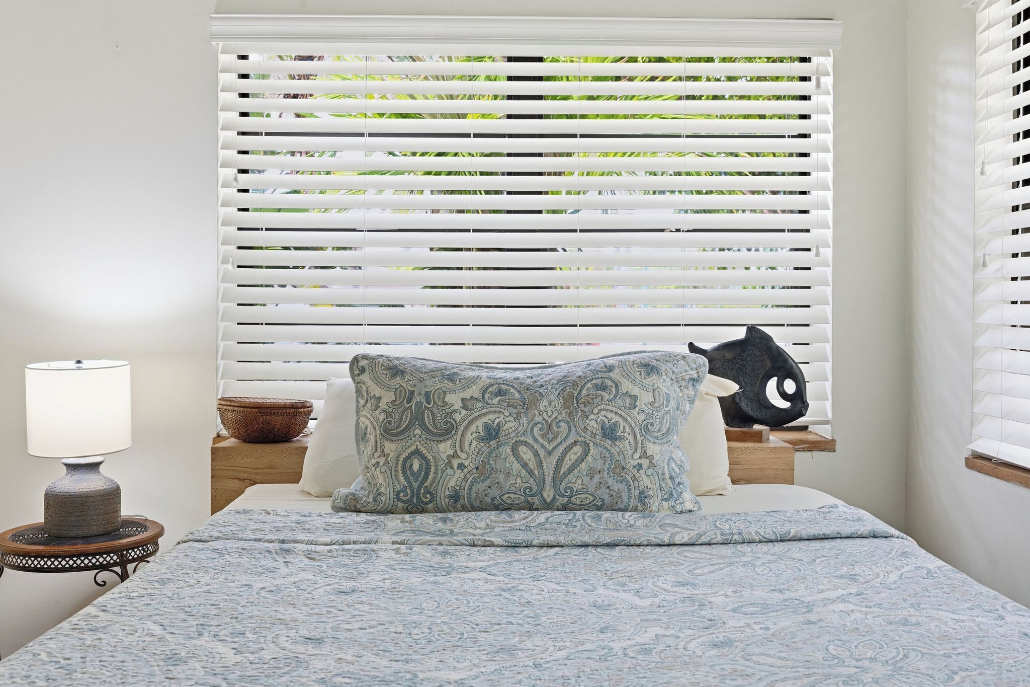 A bedroom with a bed, a bedside lamp, a decorative pillow, a small table with a bowl, and a window with blinds.