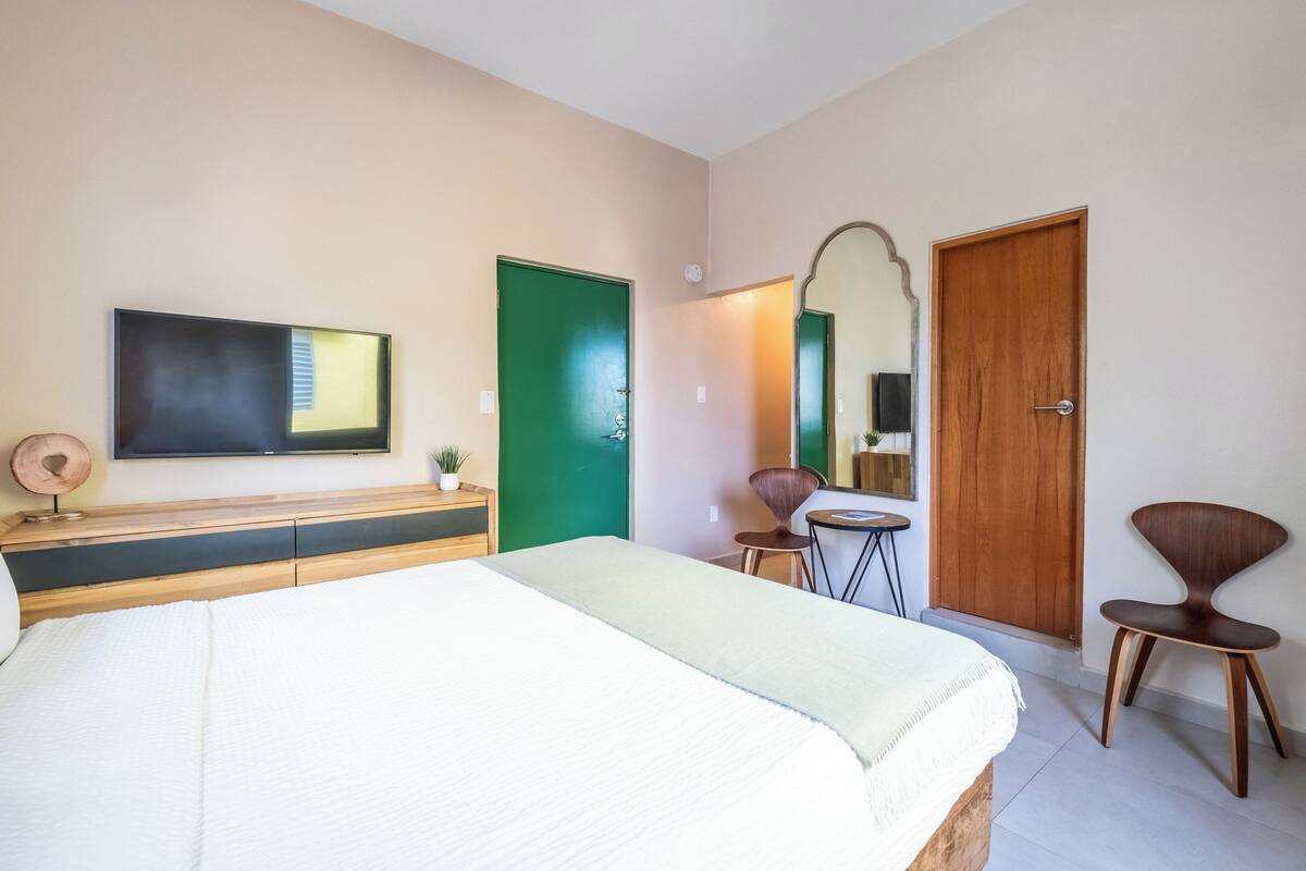 A bedroom with a bed, a TV, a green door, a wooden door, a chair, and a small table.