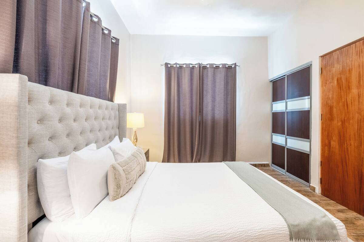 A neatly made bed with white linens and a grey headboard, a bedside lamp, and a view of a sliding door closet.