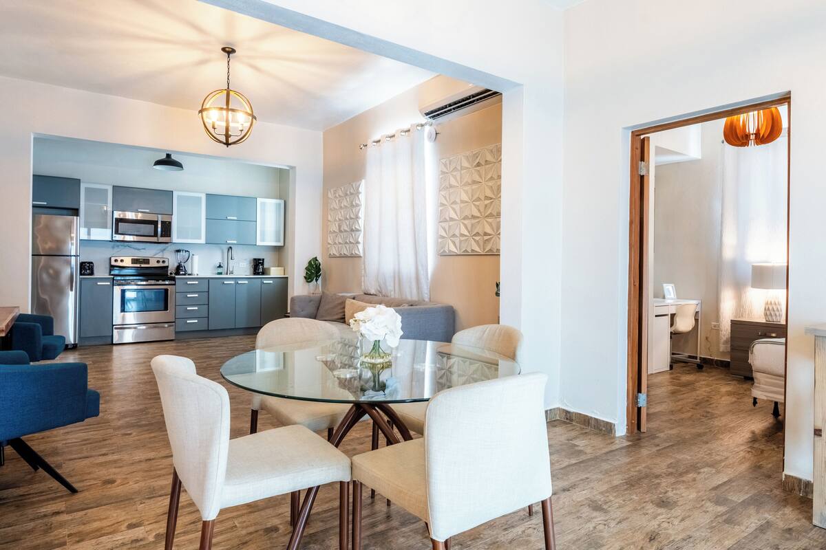 A modern kitchen with stainless steel appliances, a dining area with a glass table and white chairs, and a living area with a sofa and a decorative vase.