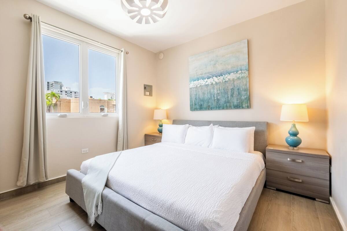 A hotel room with a large bed, two bedside tables with lamps, a window with curtains, and a painting on the wall.