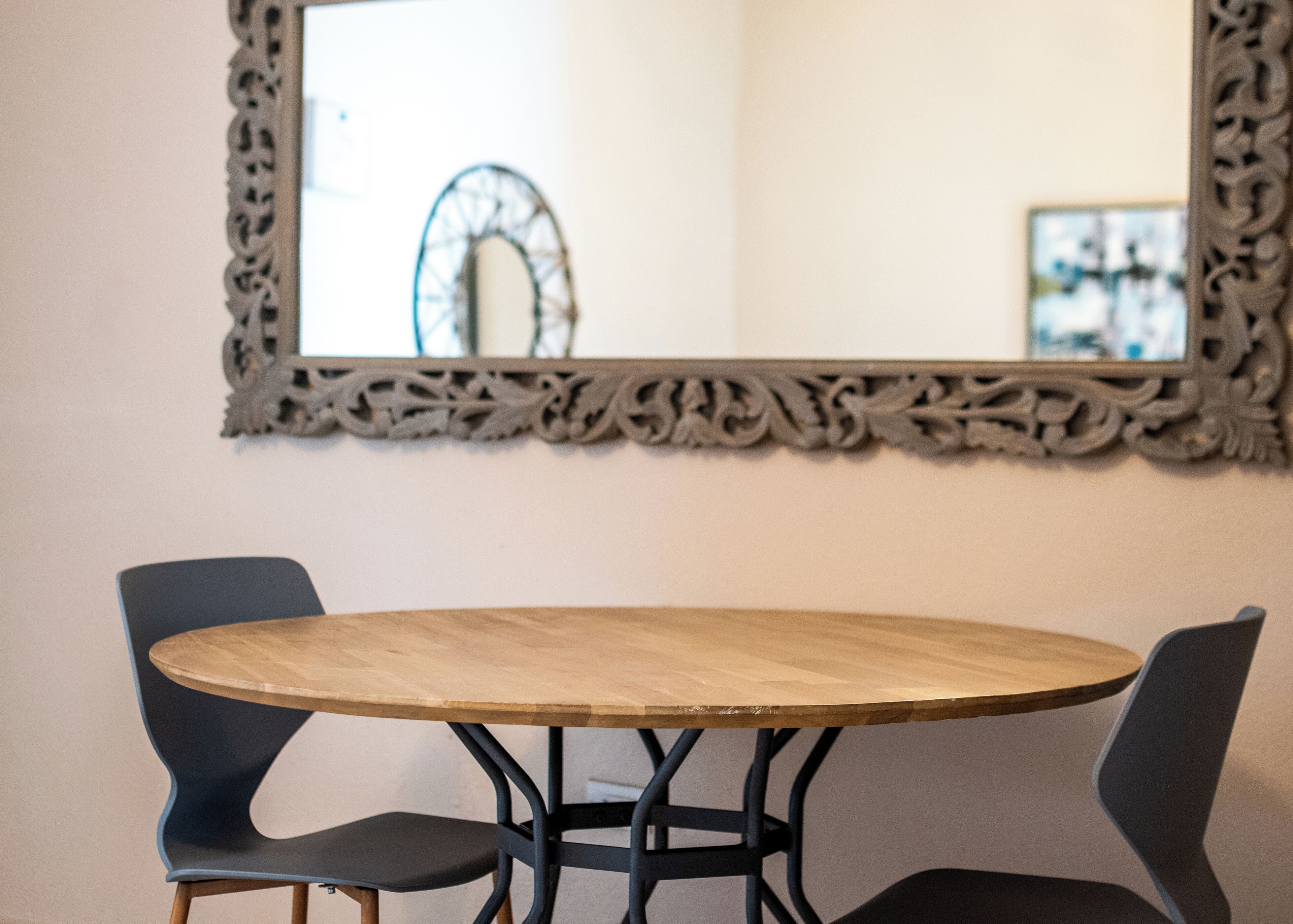 Una mesa redonda de madera con dos sillas negras, un espejo con un marco decorativo y una escultura de metal ornamental al fondo.