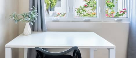 A room with a white table and a chair, a white vase with green plants, and a window with curtains.
