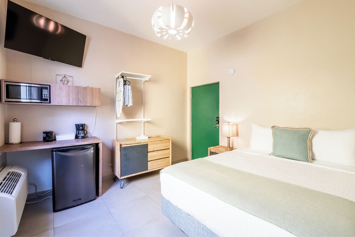 A hotel room with a bed, a desk with a microwave, a refrigerator, a TV, and a green door.