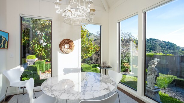 A modern dining area with a marble table, white chairs, and large windows offering a view of a garden and hills. - Sophisticated Artist's Retreat (Stinson Beach)