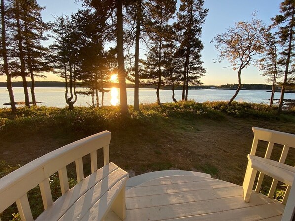 A wooden deck with two white chairs facing a lake at sunset.
