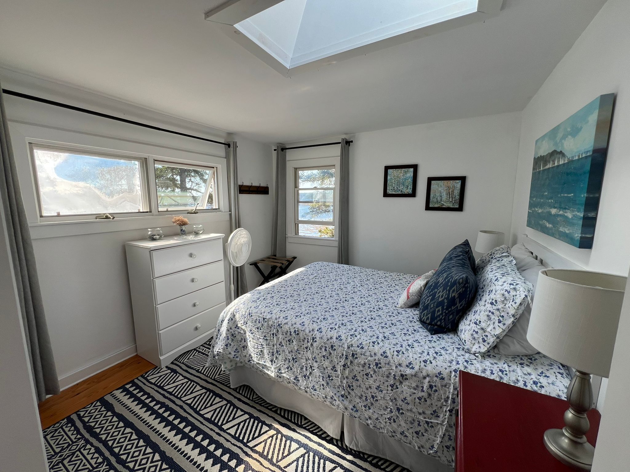 A bedroom with a skylight, two windows, a bed with floral bedding, a bedside table with a lamp, and a dresser.