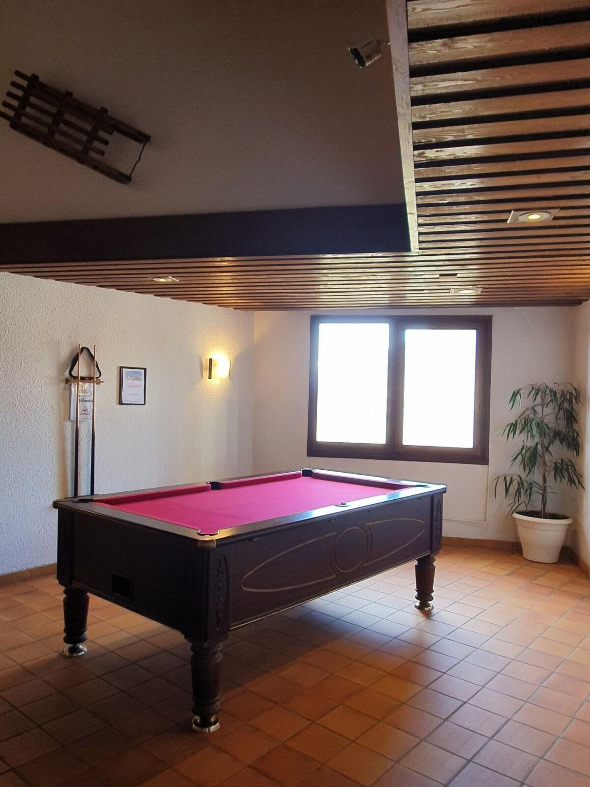 Mesa de bilhar em um ambiente com piso em mosaico, janela com persianas, planta em vaso e luminária fixada na parede.