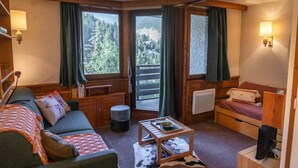Hyggelig hyttekrog med sofa, lille bord, radiator og udsigt til skov gennem store vinduer.
