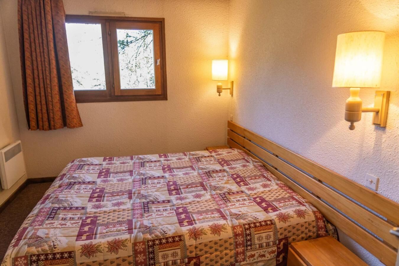 A bedroom with a bed, a window with curtains, a wall-mounted lamp, and a radiator.