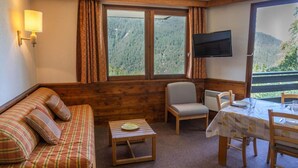 A cozy room with a wooden sofa, a small table, a chair, and a dining table with chairs. A TV is mounted on the wall, and there is a window with a view of a forested mountain. - 100m from ski slopes, 3rd floor, balcony, tv, ski locker, 39m², Les Orres (Les Orres)