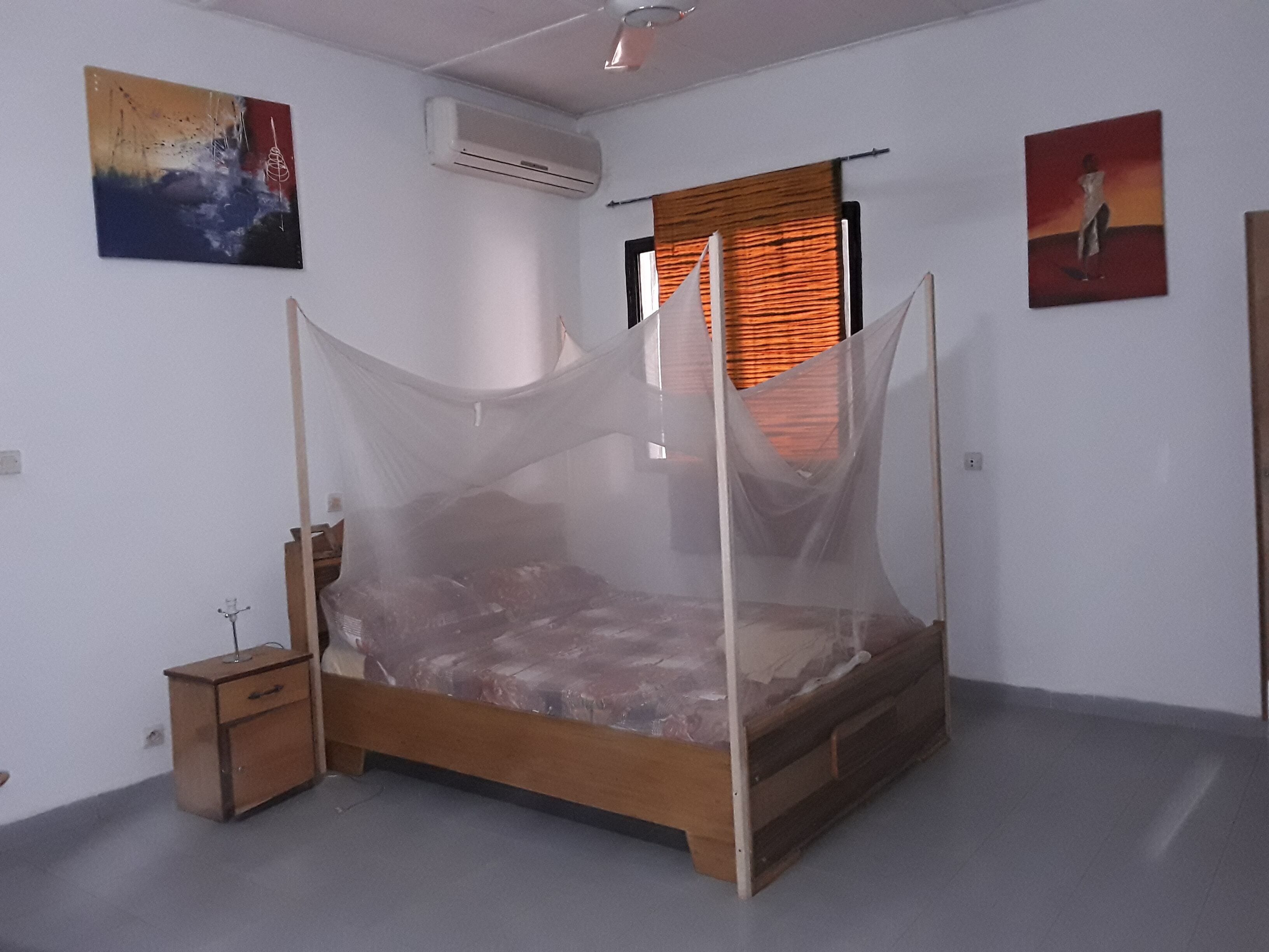 A bedroom with a wooden bed, a nightstand, a wall-mounted air conditioner, and two paintings.