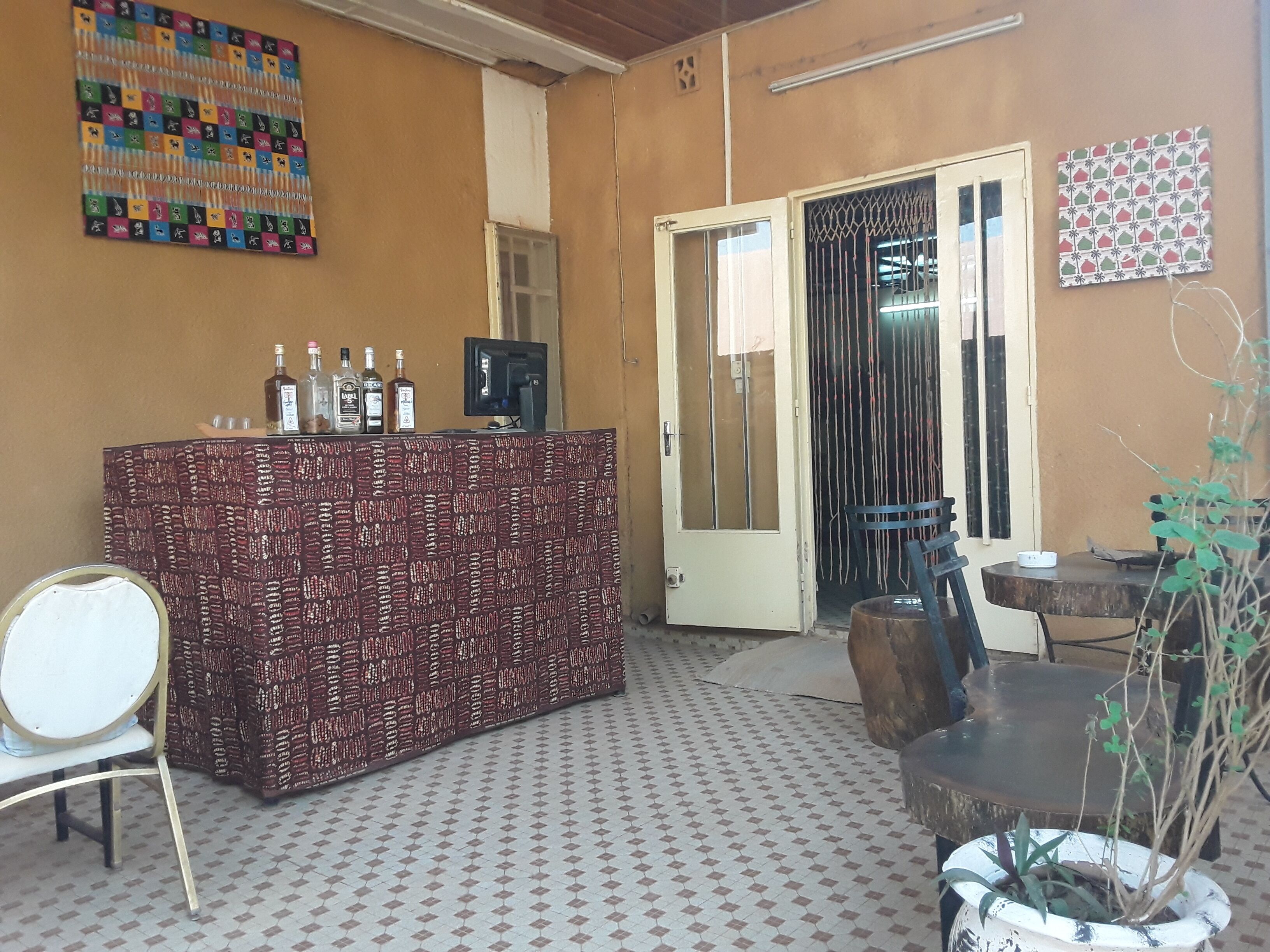 A bar area with a patterned counter, a television, and several bottles. There is a door leading to another room, a chair, and a table with a plant.