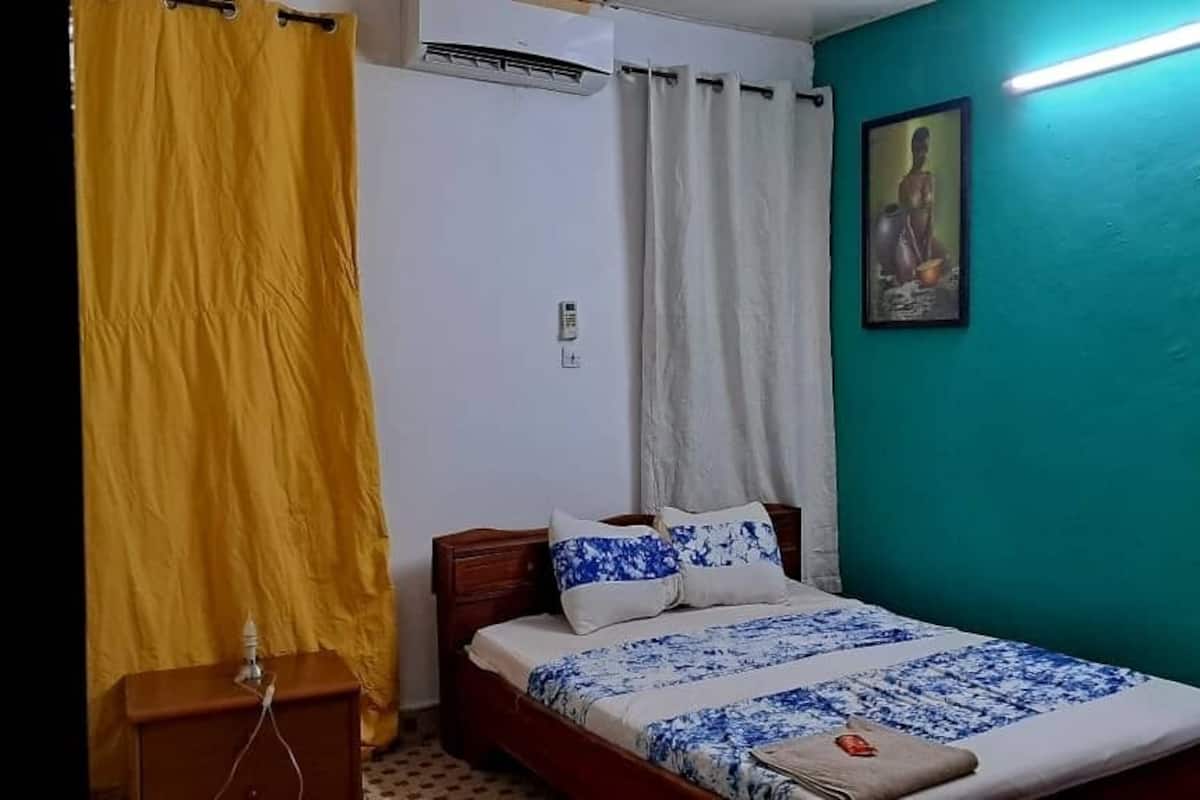 A bedroom with a bed, curtains, a wall-mounted air conditioner, a framed picture, and a bedside table.