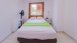 A small, clean room with a bed, a bedside table, a nightstand, a wall-mounted fan, and a window with blinds. - Apartment in Cartagena Colombia. (Cartagena)