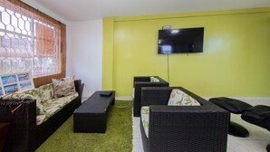 Un salon comprenant un canapé, deux fauteuils, une table basse, un téléviseur à écran plat fixé sur un mur jaune et une fenêtre avec un store en bambou.