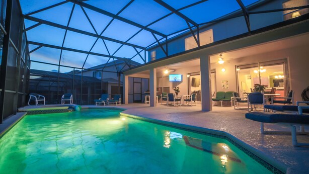 An indoor swimming pool with a glass ceiling, lounge chairs, and a television. - Enchanted Emerald Oasis by VillaDirect (Kissimmee)