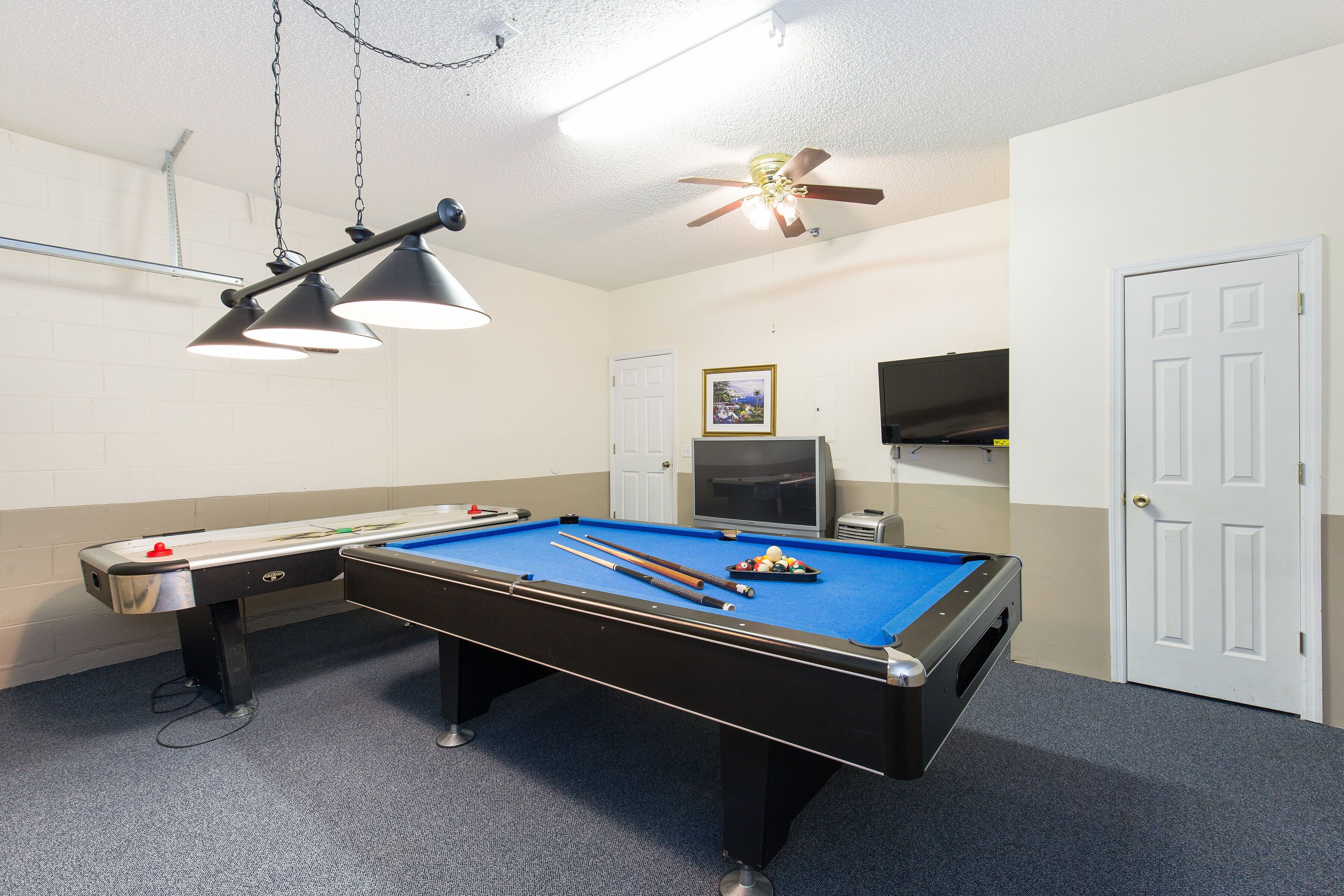 Une table de billard avec des queues et des billes, un ventilateur de plafond et un téléviseur dans une pièce aux murs blancs et au sol recouvert de moquette.
