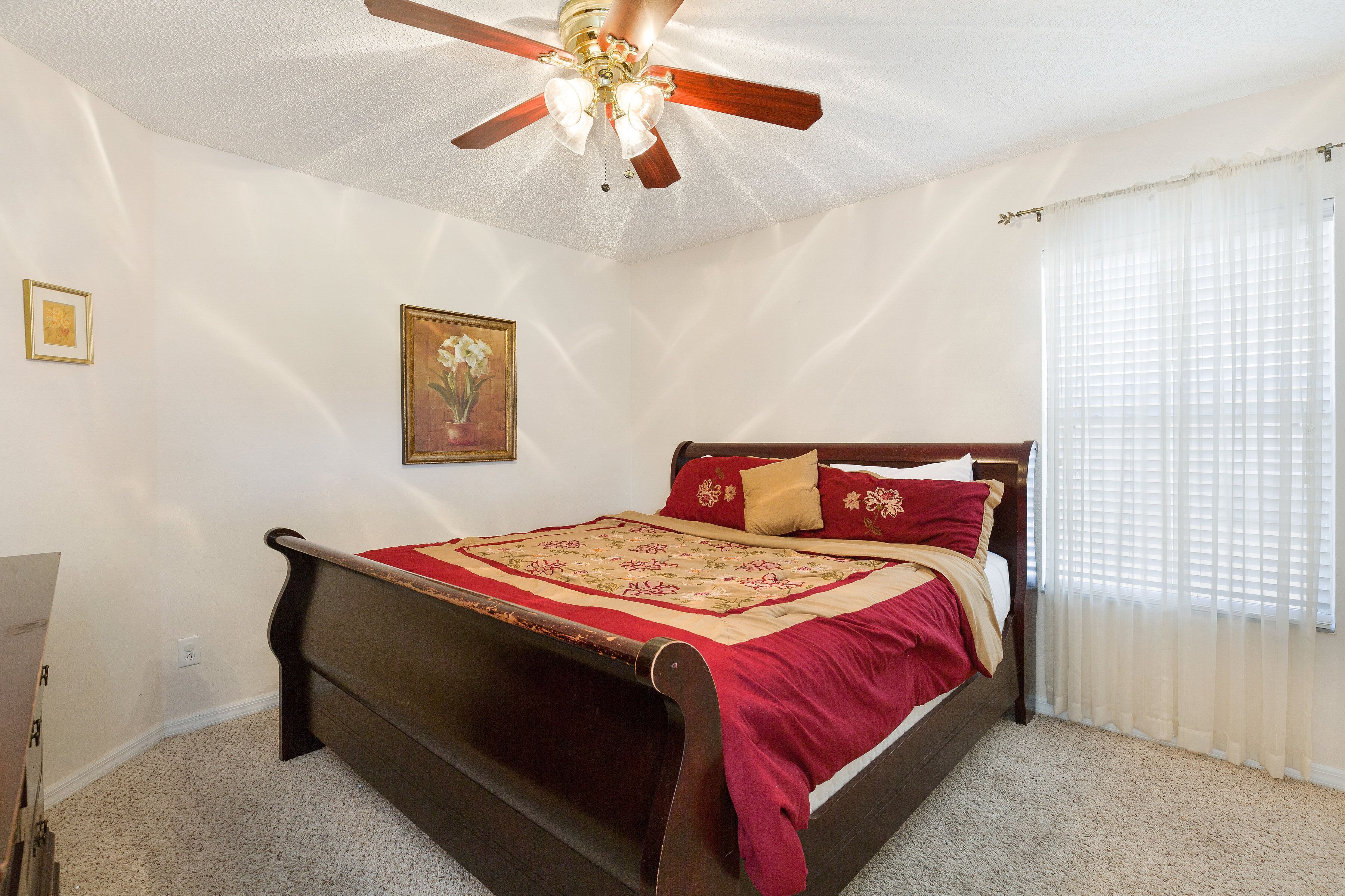 Un dormitorio con una cama de madera, un ventilador de techo, un cuadro en la pared y una ventana con cortinas.