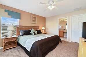A bedroom with a bed, nightstands, a ceiling fan, a window with blinds, and a door leading to a bathroom.