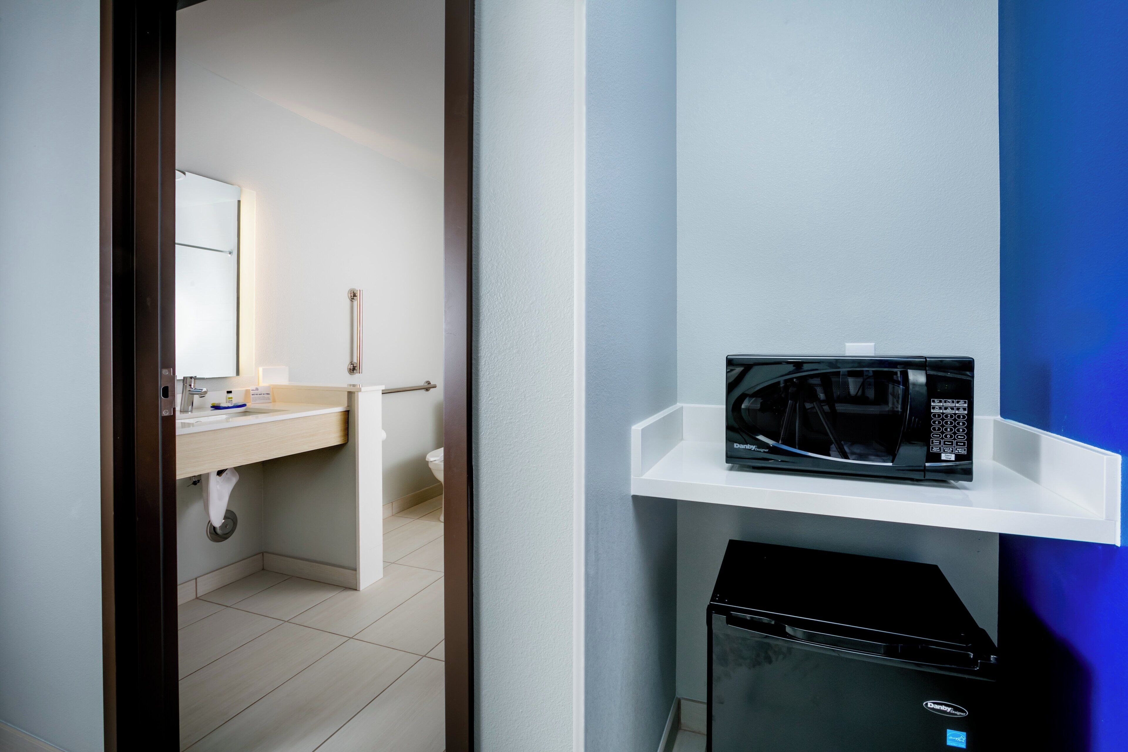Une salle de bain moderne avec un lavabo blanc, un miroir et un micro-ondes noir posé sur une étagère blanche.