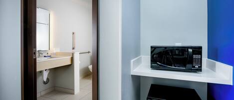 A modern bathroom with a white sink, mirror, and a black microwave on a white shelf.