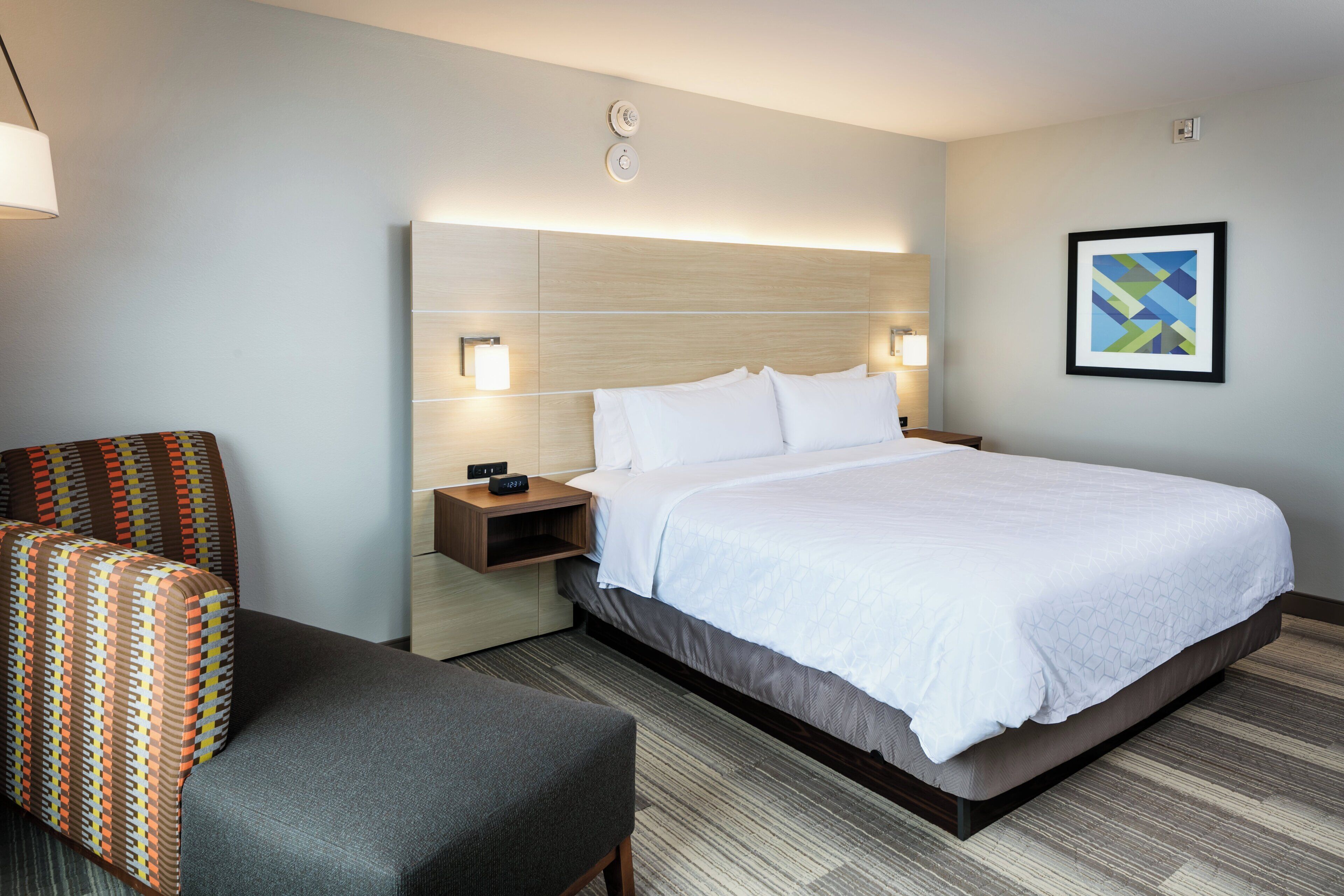 Una habitación de hotel con cama, una silla, mesitas de noche y una lámpara fijada a la pared.