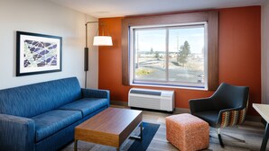 A modern living room with a blue sofa, a wooden coffee table, a patterned ottoman, and a black armchair. There is a large window with a view of a cityscape. - Holiday Inn Express & Suites Bend South by IHG (Bend)
