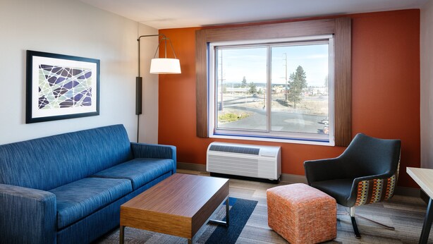 A modern living room with a blue sofa, a wooden coffee table, a patterned ottoman, and a black armchair. There is a large window with a view of a cityscape. - Holiday Inn Express & Suites Bend South by IHG (Bend)