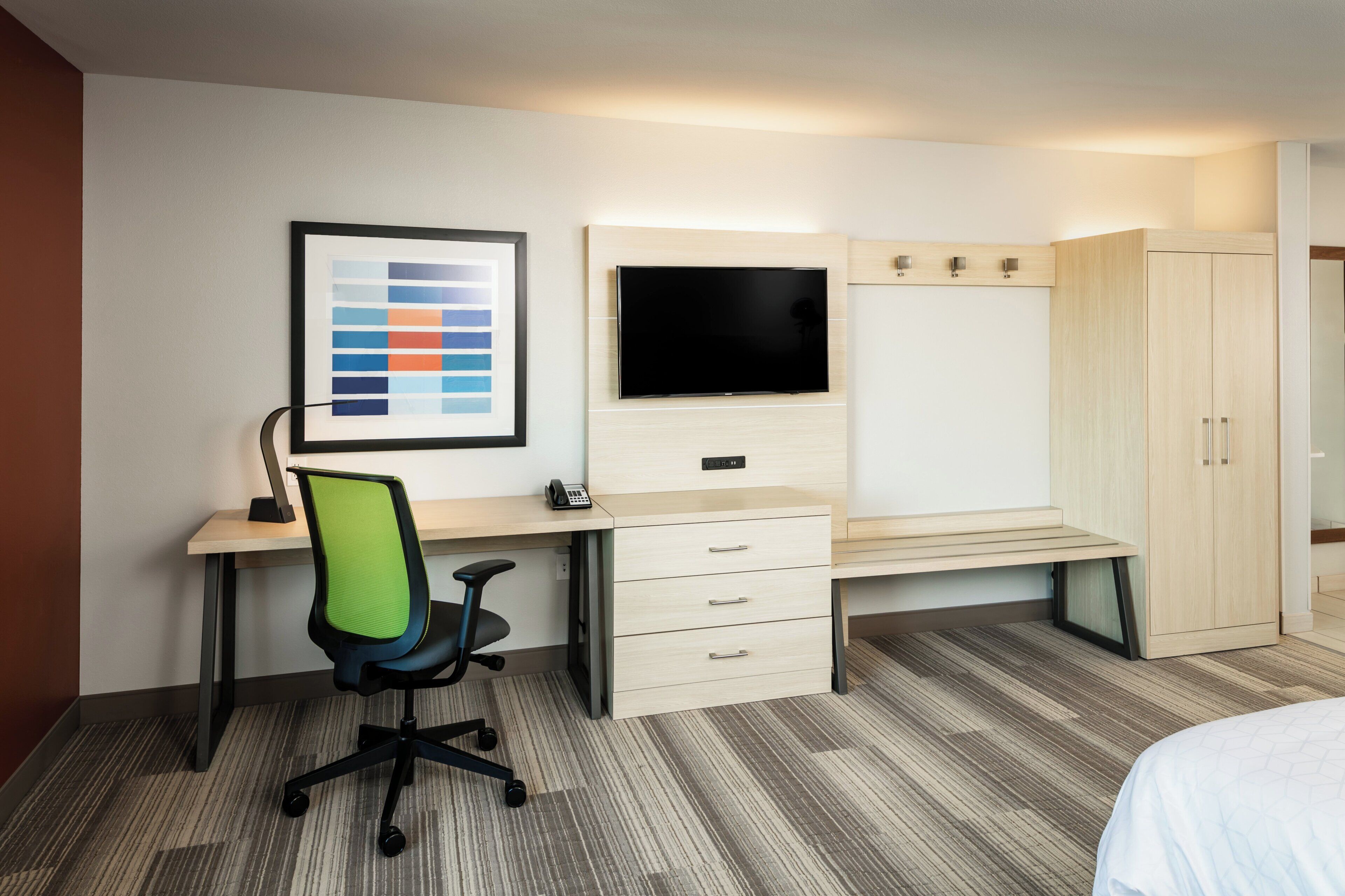 A hotel room with a desk, chair, wardrobe, and a flat-screen TV.