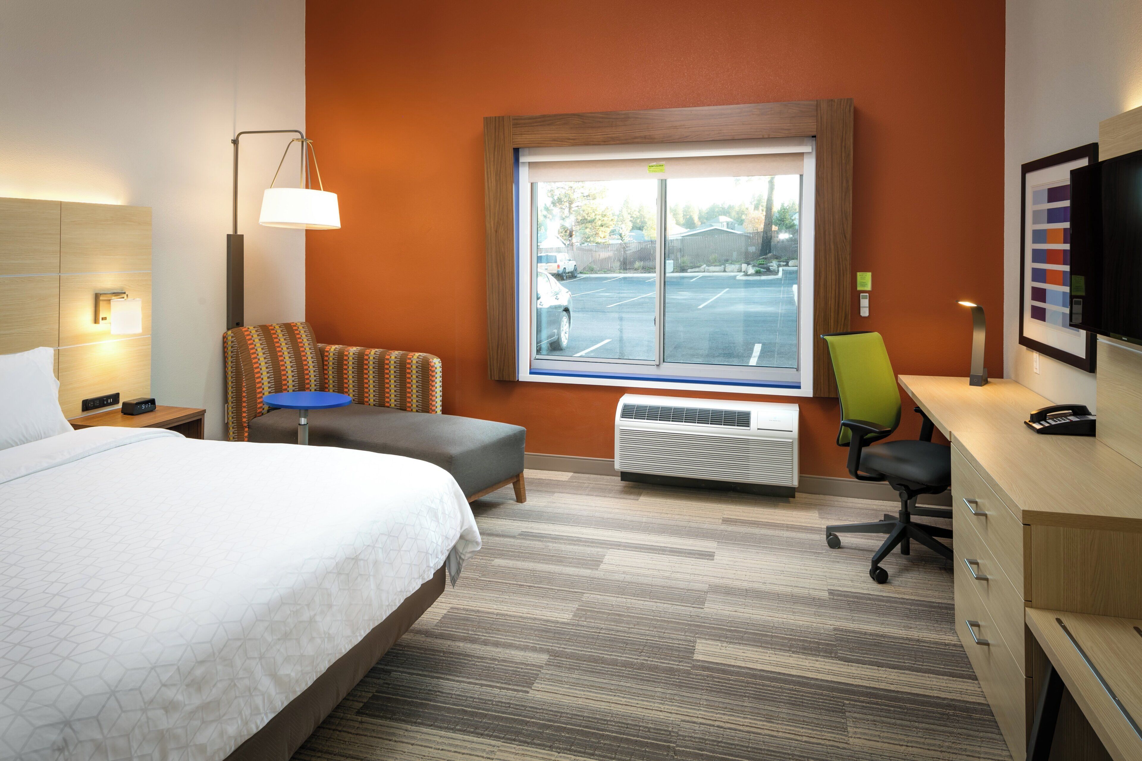 Ein Hotelzimmer mit einem Bett, einem Schreibtisch, einem Stuhl, einem Heizkörper und einem Fenster mit Blick auf einen Parkplatz.