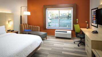 Habitación de hotel con cama, escritorio, silla, calefactor y ventana con vista a un estacionamiento.