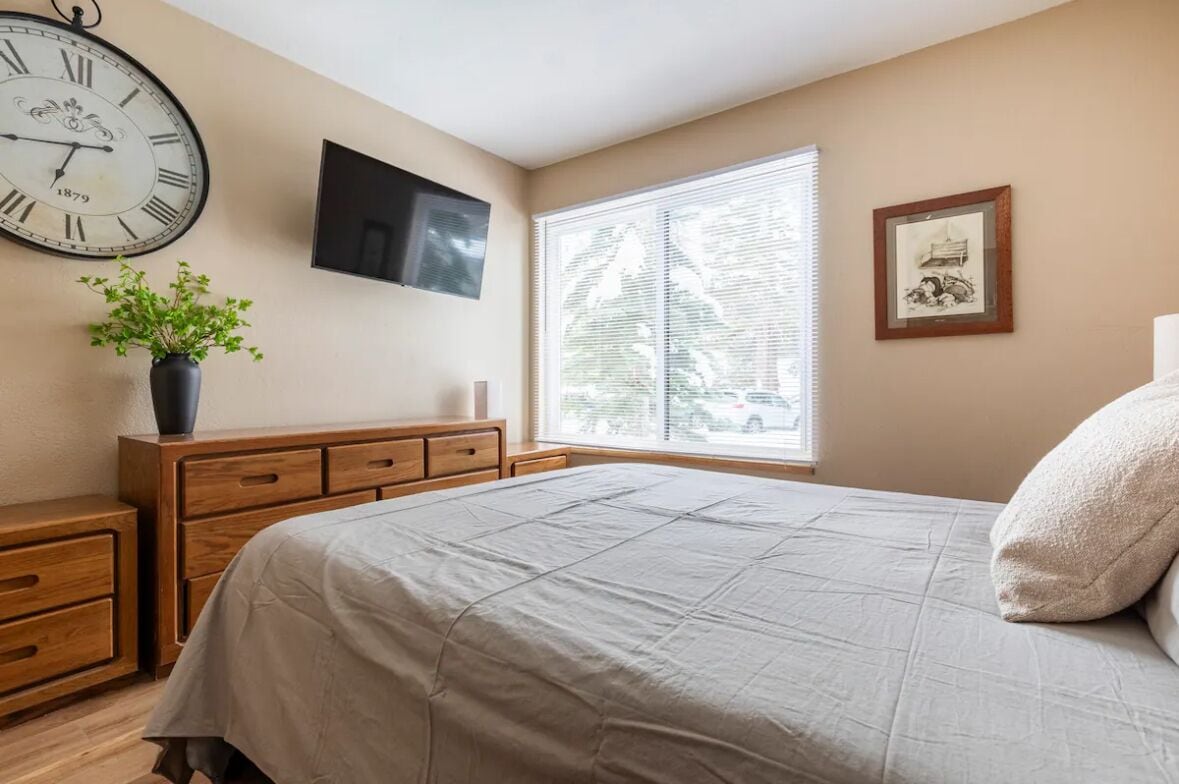 A bedroom with a bed, a wooden dresser, a clock, a television, a window with blinds, and a framed picture on the wall.