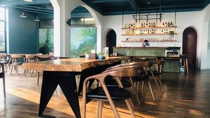 A modern restaurant interior with wooden tables and chairs, a bar area with shelves of bottles, and a large window with a view of greenery. - Le Vent Homestay Tam Dao (Tam Dao)