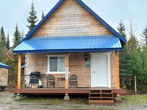 A wooden cabin with a blue roof, a front door, and a small porch with two chairs.