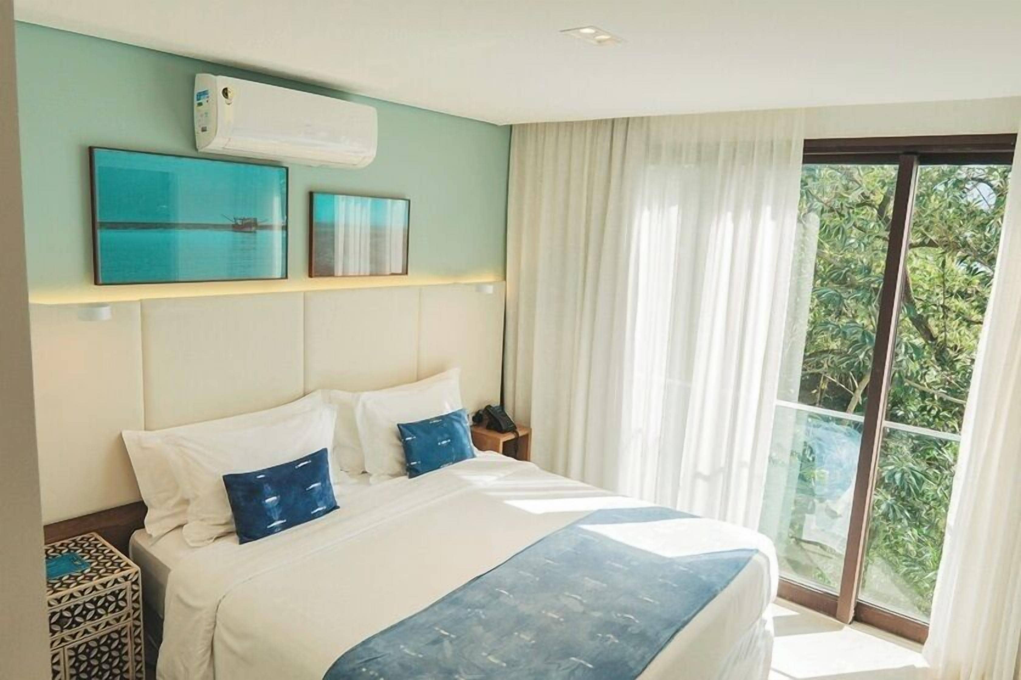 A hotel room with a bed, a nightstand, a wall-mounted air conditioner, and a large window with curtains.