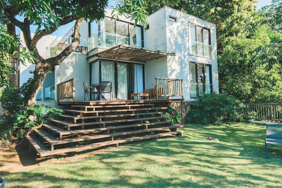 Una casa moderna de dos pisos con un porche de madera, un área de comedor con sillas y un árbol grande al frente.