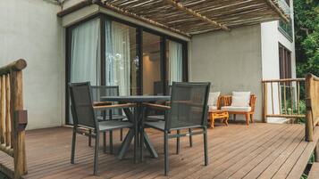 Um deck de madeira com mesa e cadeiras, uma área coberta com telhado de bambu e vista para o verde.