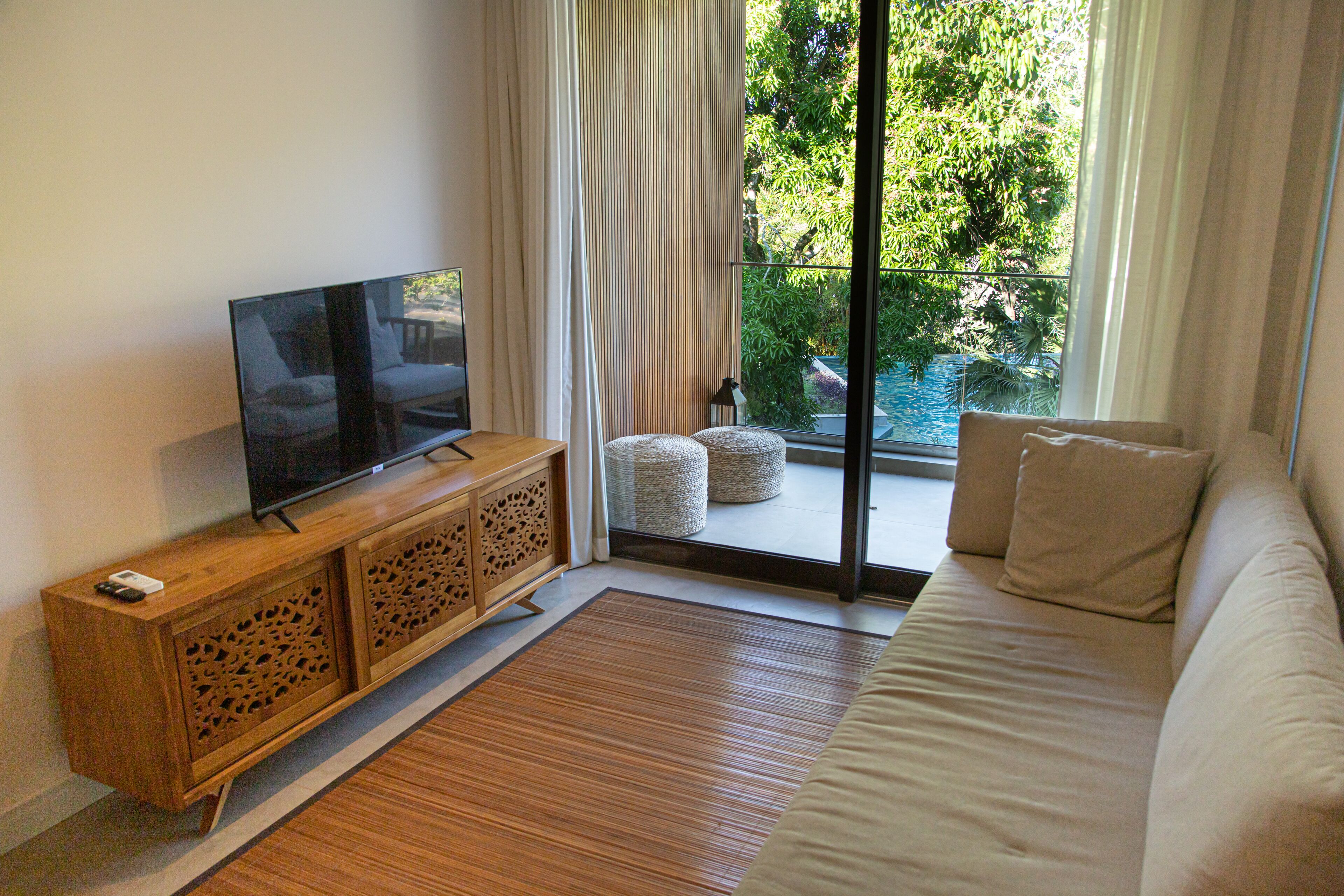 A living room with a TV on a wooden cabinet, a beige sofa, and a view of greenery through a glass door.