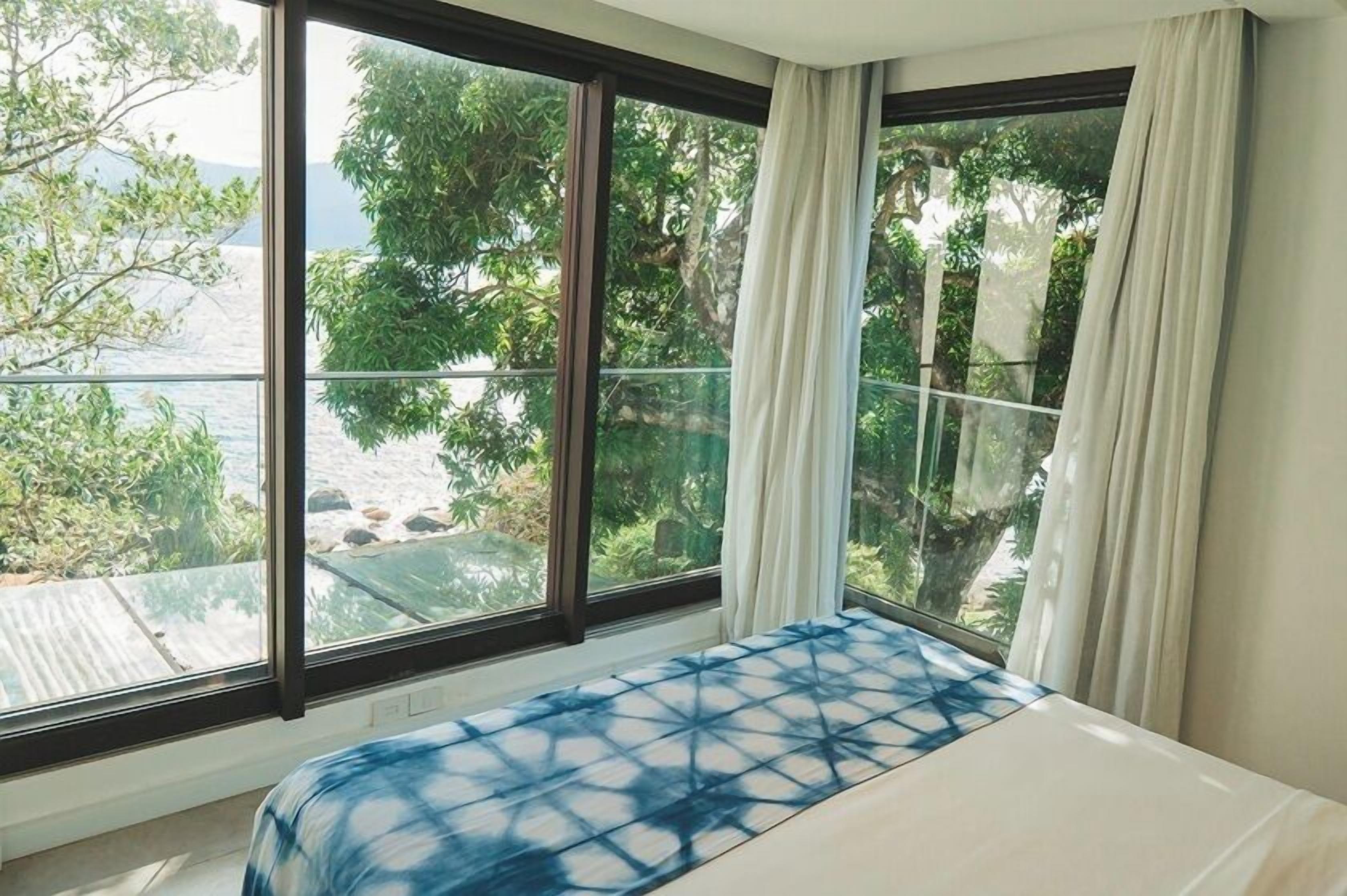 A bedroom with a large window, a bed with a blue patterned bedspread, and a view of a beach and trees.