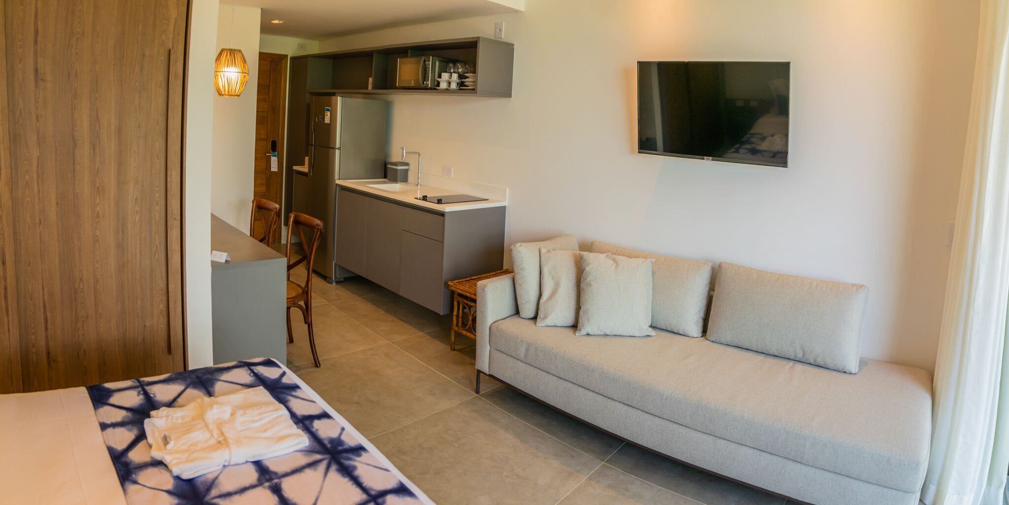 Una habitación de hotel compacta con cama, sofá, kitchenette pequeño y televisor montado en la pared.