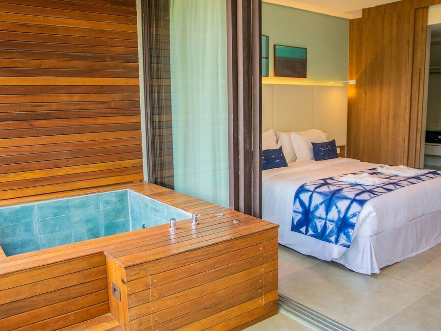 Habitación de hotel con pared de paneles de madera, una bañera grande de madera, una cama con colcha azul estampada y vistas a un paisaje urbano a través de una ventana.