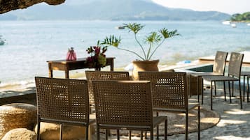 Relax in a serene outdoor setting with wicker chairs and a table, surrounded by a calm sea and distant mountains.