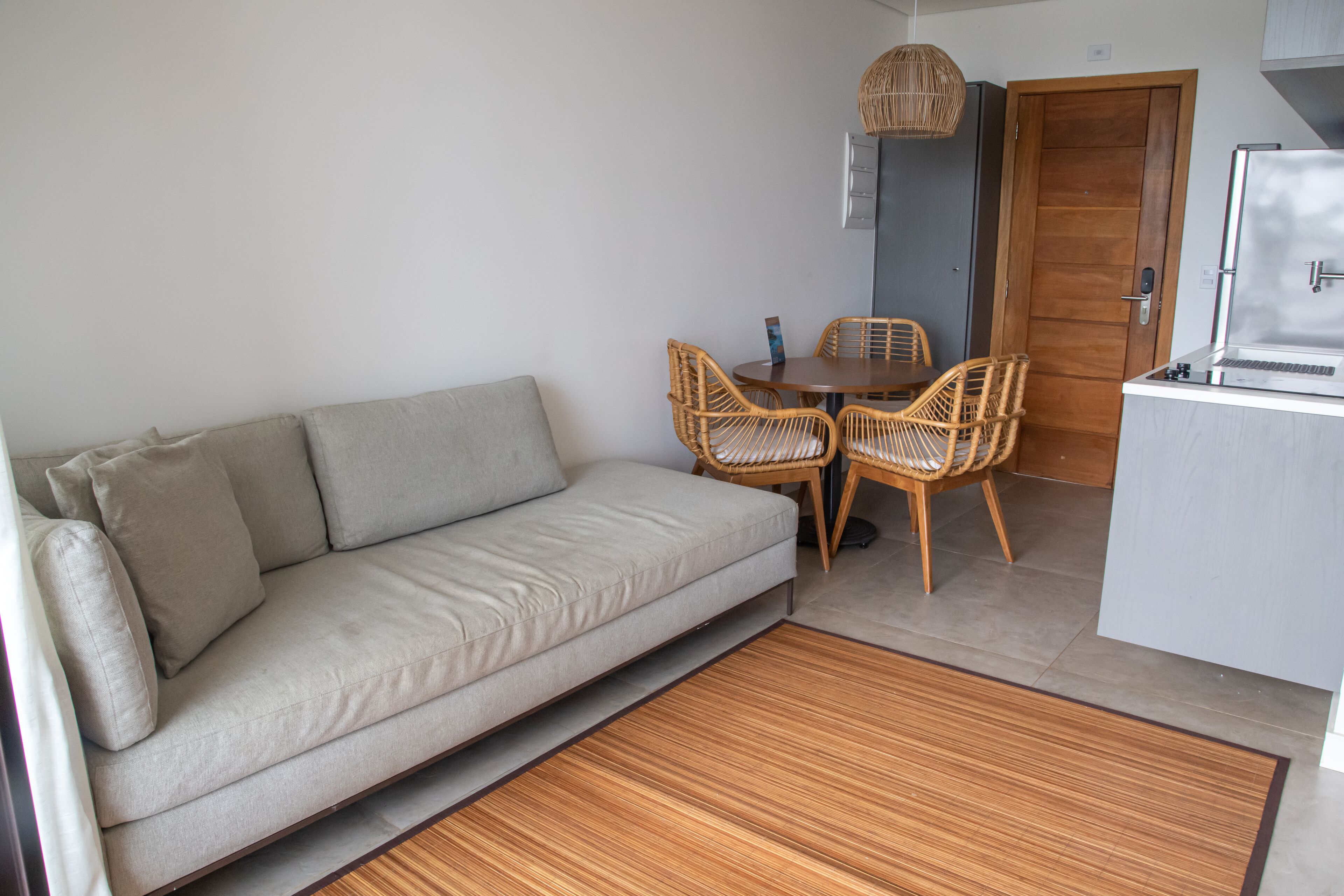 A modern living room with a sofa, a dining table with chairs, a kitchen area with a refrigerator, and a wooden floor.