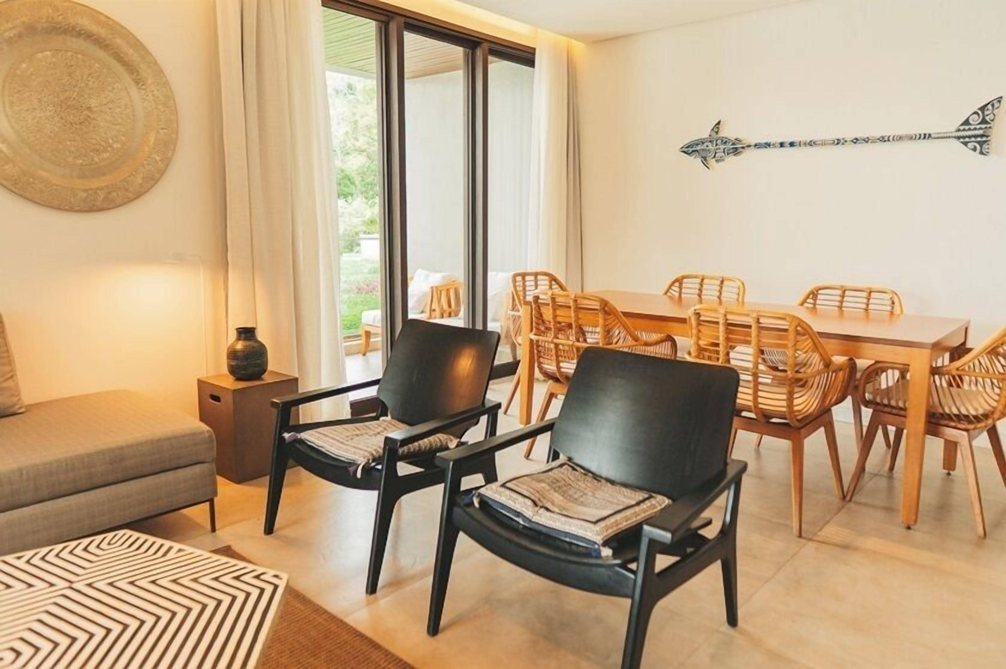 Un salon moderne comprenant un canapé, deux fauteuils noirs, une table à manger en bois avec six chaises, et une grande fenêtre avec des rideaux.