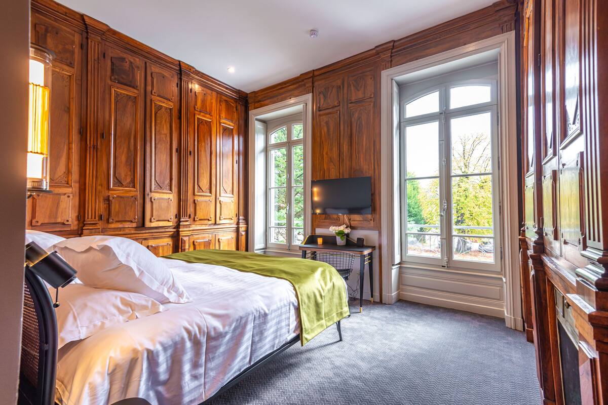 Ein Schlafzimmer mit Holzwänden, einem Bett, einem Fernseher und einem Fenster mit Blick ins Grüne.