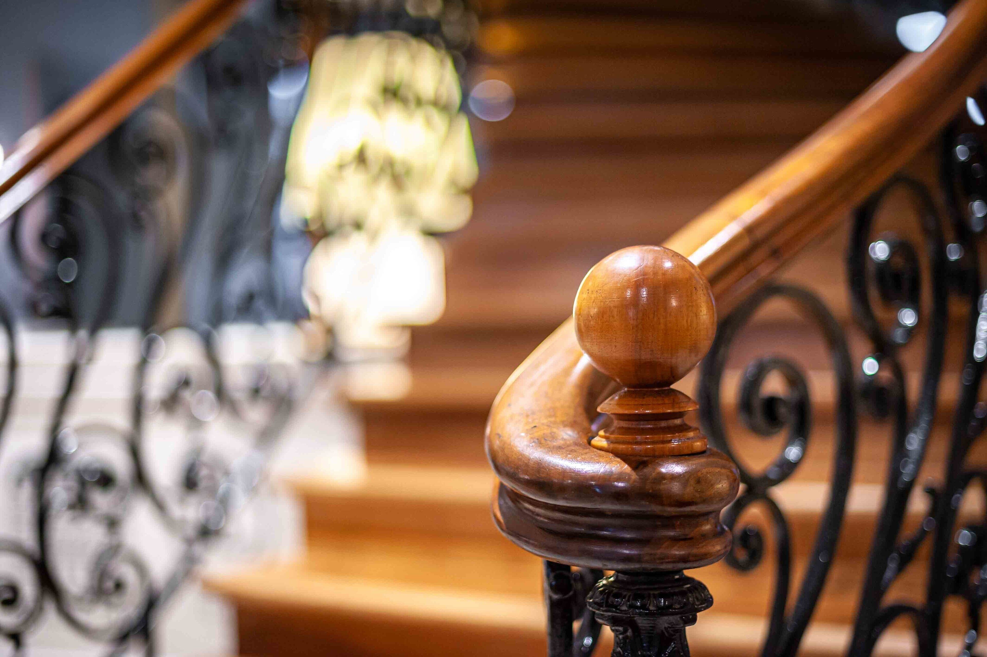 Een houten leuning met een ronde knop, deel van een trap met versierde zwarte metalen spijlen.