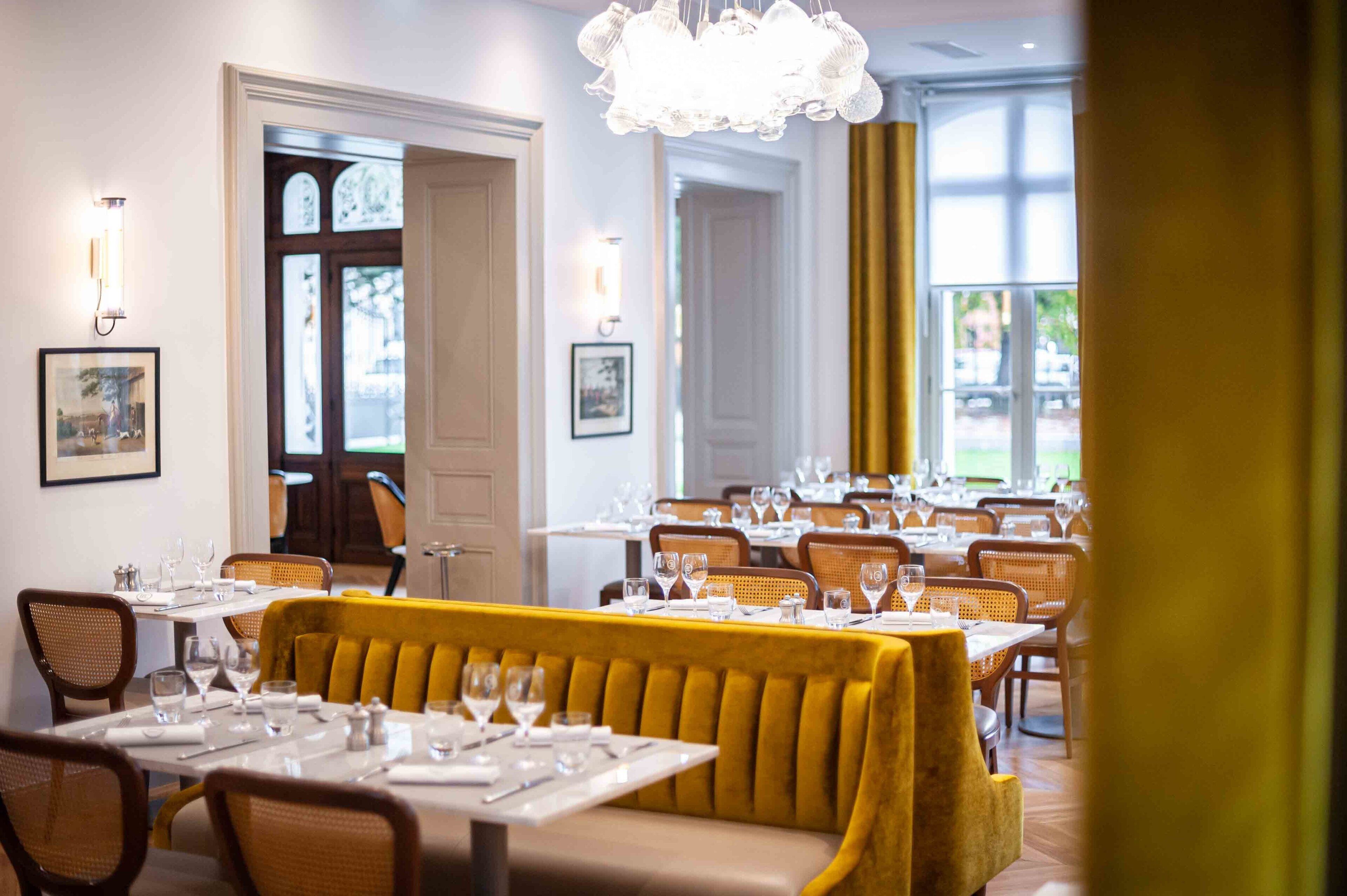 A restaurant with a yellow upholstered bench, white table settings, wicker chairs, and a chandelier.